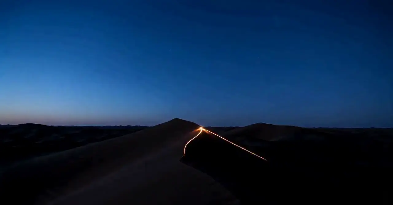 dark desert dune ridges at blue hour, most lying low while a sparse few rise tall and catch a warm champagne light along their crests, one glowing warm orange